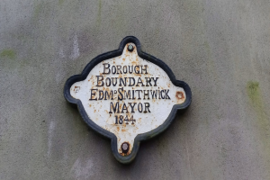 Metallplatte an einer Gebäudeseite, die die Borough-Grenze markiert, mit "Borough Boundary Edm Smithwick Mayor" in fetten weißen Buchstaben auf schwarzem Hintergrund.