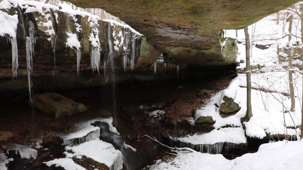 Ein kleiner Wasserfall ergäßt sich an einem schneebedeckten, steinigen Hang in einem wöldernen Gebiet, mit Eiszapfen an den Steinen und schneebedeckten Bäumen im Hintergrund.