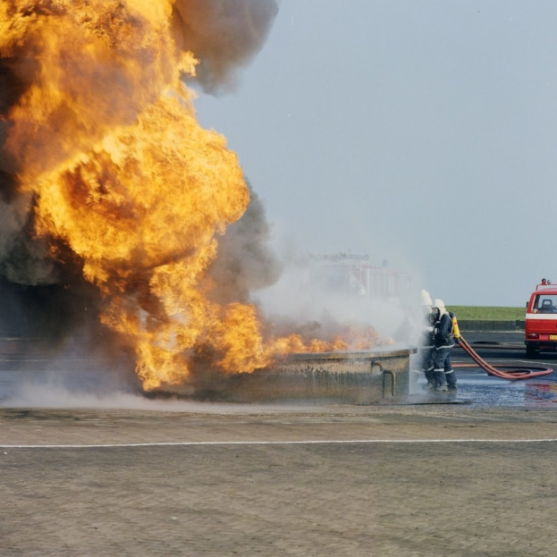 Feuerwehrlöschfahrzeug von Flammen umgeben auf der Straßenseite mit zwei Helmen tragenden Individuen, die Rohre halten, einem Fahrzeug im Hintergrund und dem Himmel.