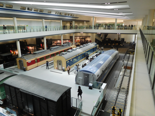 Ein geräumiger Museumsraum voller einer bunten Sammlung von Zügen, die von Besuchern betrachtet werden, mit Geländern, Säulen und Deckenleuchten.