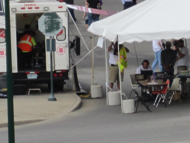 Ein Krankenwagen steht links von einer StraÃŸe, wÃ¤hrend auf der rechten Seite ein Zelt eine Gruppe von Menschen schÃ¼tzt, mit einem Tisch und Stuhl in der NÃ¤he.