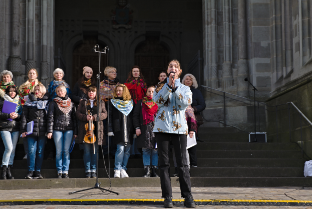 Eine Gruppe von Menschen vor einem Gebäude, mit einer Frau in der Mitte, die ein Mikrofon hält und singt, umgeben von Mikrofonen mit Ständern, Musikinstrumenten, Treppen mit Geländern, einer Wand mit Säulen und einer Tür auf der rechten Seite.