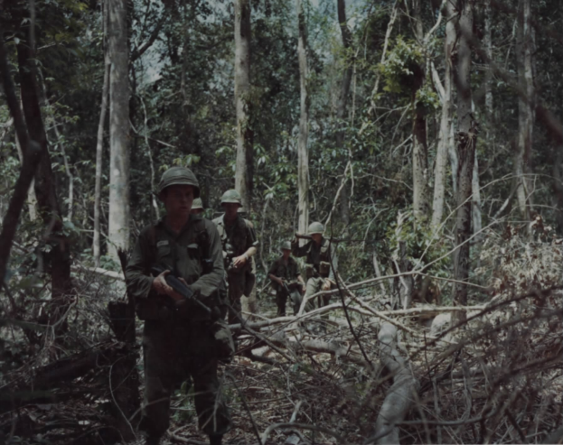 Eine Gruppe von Soldaten in Helmen und mit Gewehren durchquert einen grünen Wald mit hohen Bäumen, mit Text am unteren Bildrand.