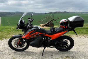 Eine KTM 1290 Super Adventure R Motorrad auf einem Schotterweg mit Helm und Gepäckstück daneben, umgeben von grünem Gras, hohen Bäumen und Strommästen unter einem bewölkten Himmel.