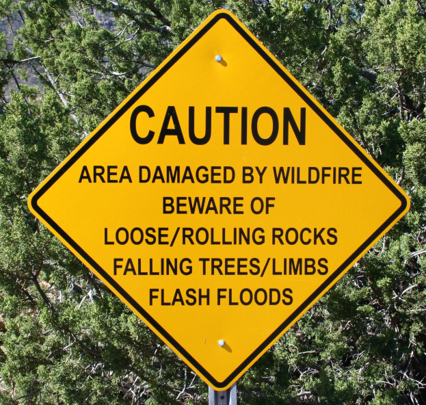 Gelbes Warnschild an einem Pfosten in der Nähe, das vor Waldbrandschäden, lockeren Steinen, umstürzenden Bäumen und Überschwemmungen warnt, mit Bäumen im Hintergrund.