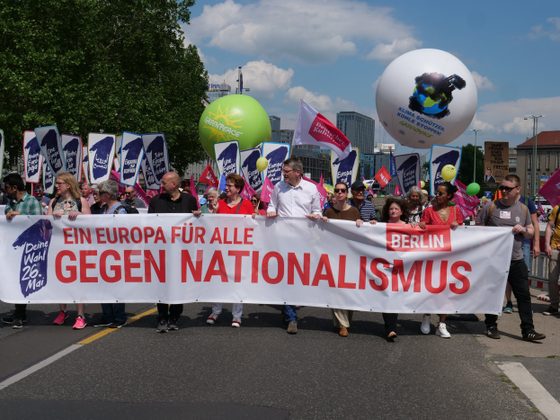 Eine Gruppe von Menschen marschiert bei einer Demonstration gegen Nationalismus in Berlin und trägt ein Banner, Fahnen und Luftballons bei sich, während im Hintergrund Bäume, Gebäude und ein bewölkter Himmel zu sehen sind.