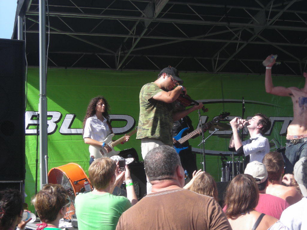 Eine Gruppe von Musikern, die verschiedene Instrumente auf einer Bühne spielen, mit einem großen schwarzen Lautsprecher links daneben, während das Publikum davor steht und die Performance genießt, mit einem grünen Banner im Hintergrund.