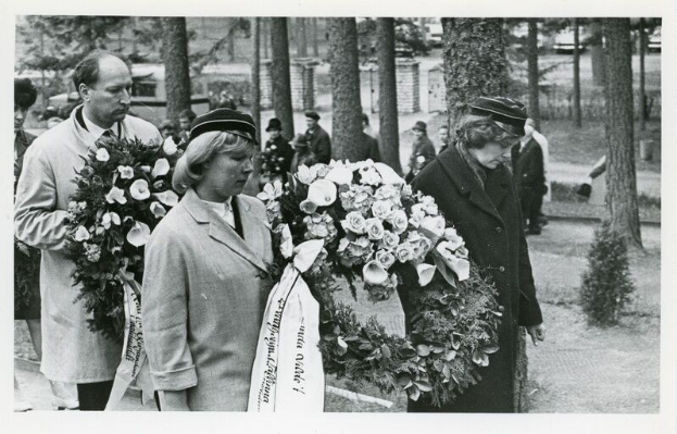 Schwarz-weißes Bild einer Gruppe von Menschen, die Blumensträuße bei einer Gedenkfeier für Holocaust-Opfer halten, mit Bäumen und Gebäuden im Hintergrund.