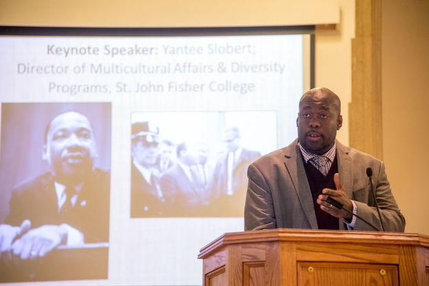 Ein Mann in Mantel, Krawatte und Hemd spricht rechts neben einem Rednerpult, mit einem Projektionsschirm in der Mitte und einem Mann in Schwarz-Weiß links.