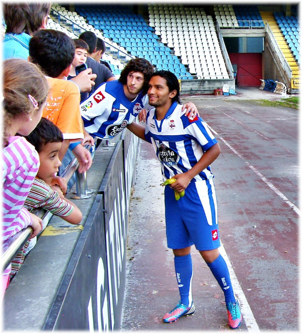 Ein Sportler steht in der Mitte eines Bildes mit leeren Stühlen drumherum, während das Publikum auf der linken Seite hinter einer Begrenzung steht und ein Hintergrundbereich sichtbar ist.