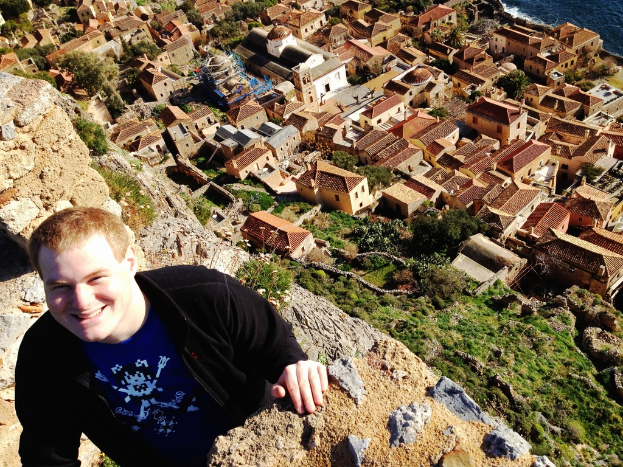 Ein Mann in einer schwarzen Jacke und einem blauen T-Shirt steht lachend auf der linken Seite des Bildes, mit H├Ąusern und B├Ąumen im Hintergrund.