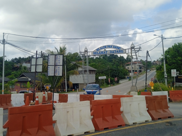 Ein Auto fährt auf einer Straße mit orange-weißen Barrieren, Verkehrskegeln, Laternenmasten, Strommasten mit Drähten, einem Bogen mit Text, Gebäuden mit Fenstern, Bäumen und einem bewölkten Himmel im Hintergrund.