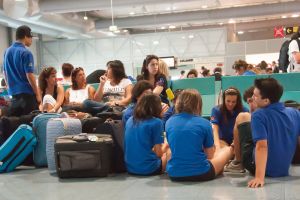 Ein belebtes Flughafeninnere mit vielen Menschen, die auf Flüge warten, Gepäck auf der linken Seite und Glastüren mit Hinweisen im Hintergrund.