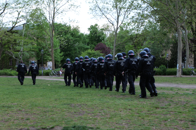 Gruppe von Polizisten in schwarzen Uniformen und Helmen, die über ein grünes Feld gehen, mit Fahrrädern, Laternenmasten, Pflanzen, Bäumen, Gebäuden und einem klaren blauen Himmel im Hintergrund.