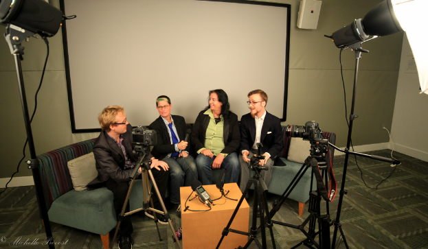 Eine Gruppe von drei Personen sitzt auf einem Sofa und gibt ein Interview. Links befindet sich eine Person in einem schwarzen Mantel und Hose, in der Mitte eine Person in einem Mantel und blauer Krawatte, die ein Mikrofon hält, und rechts eine strahlende Person. Auf dem Tisch in der Mitte steht ein Mikrofonrecorder, Kameras sind auf der rechten und linken Seite und große Scheinwerfer sind vorhanden, im Hintergrund ein Projektionsschirm.