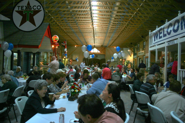 Menschen sitzen an Tischen mit Getränkedosen, Servierplatten, Blumentöpfen und Papiertischdecken, während andere in der Nähe stehen; im Hintergrund sind Namensschilder, Luftballons, Elektroleuchten, Wandschmuck und Dekoleuchten an Pflanzen zu sehen.