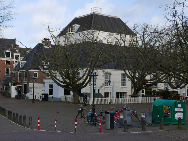 Gruppe von Fahrrädern vor einem Rathausesgebäude geparkt, mit Menschen in der Nähe, umgeben von Bäumen, Pfählen, einem Zaun und Gebäuden unter einem bewölkten Himmel.
