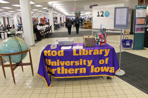 Eine Ausstellung mit einem Tisch, auf dem verschiedene Landesflaggen, eine Box, Papiere und ein Banner sowie links daneben eine Weltkugel, rechts eine Mitteilungstafel mit einigen Menschen in der Nähe und mehrere Tische und Stühle im Hintergrund zu sehen sind.