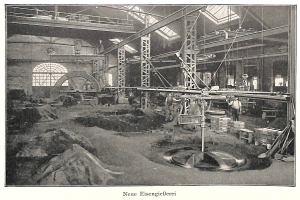 Schwarzes und weißes Foto einer Fabrik mit Menschen, die an einer Maschine arbeiten, umgeben von Metallstäben und -pfosten, beschriftet mit "Neue Eisenglebeerei, die erste Fabrik Deutschlands" unten.