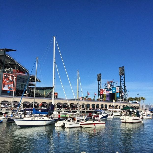 Boote auf dem Wasser im Vordergrund mit einer Brücke, Bögen, Fahnen, Bannern, Gebäuden, Pfählen, Bäumen und einem bewölkten Himmel im Hintergrund.