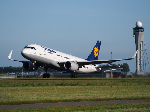 Ein Airbus A320-200 der Lufthansa startet am Frankfurter Flughafen, mit grünem Gras und einer Straße im Vordergrund und hohen Türmen, Bäumen und einem klaren blauen Himmel im Hintergrund.