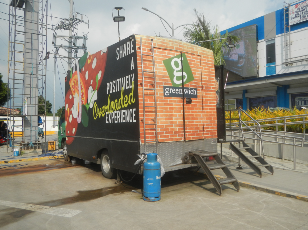 Ein Lastwagen steht vor einem Gebäude mit einer Tafel, auf der "Teilen Sie ein positiv überladenes Erlebnis" steht, mit Stufen, die zum Lastwagen führen, einem Zylinder auf dem Boden, Geländern, Strommasten mit Drähten, Laternenmasten, Bäumen und einem bewölkten Himmel im Hintergrund.
