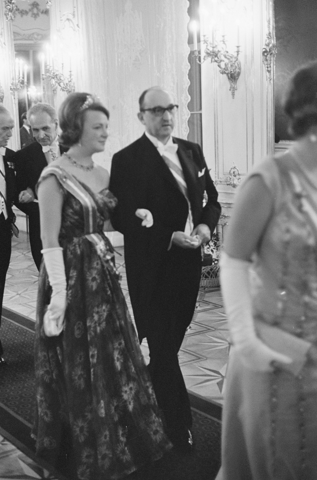 Ein Mann in einem schwarzen Blazer, weißem Hemd und schwarzen Hosen sowie eine Frau in einem eleganten Kleid gehen einen Flur entlang, umgeben von einer Gruppe von Menschen, die auf dem Boden stehen, während ein paar an der Wand montierte Lampen eine gemütliche Atmosphäre schaffen.