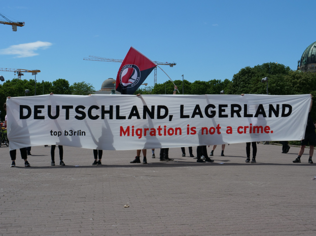 Eine Gruppe von Menschen mit einem Transparent mit der Aufschrift "Deutschland, Lagerland Migration ist kein Verbrechen" und einer Flagge, im Hintergrund Straßenmäste, Straßenlaternen, Bäume, Kräne, ein Gebäude und ein bewölktes Himmel