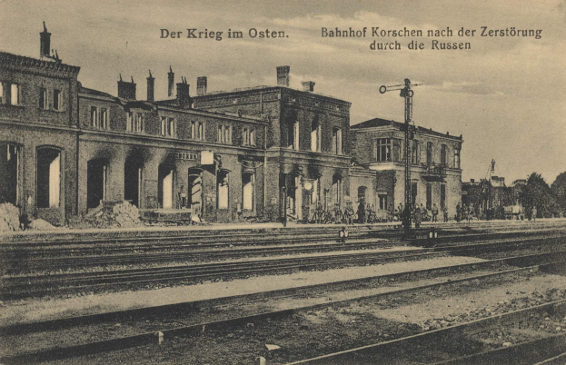 Schwarzes und weißes Foto des Bahnhofs Korschen in Ostern, Deutschland, zeigt Bahnschienen, Bahnhofsgebäude, Menschen, Bäume und einen Himmel mit dem Text "Bahnhof Korschen in Ostern, Deutschland" oben und unten.