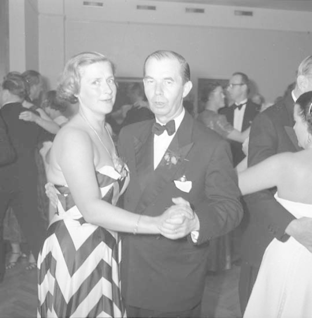 Ein Mann in einem schwarzen Blazer, weißem Hemd und schwarzen Hosen tanzt mit einer Frau in einem unbekannten Outfit auf einer formellen Veranstaltung, indem sie sich an den Händen halten und zur Musik schwenken.