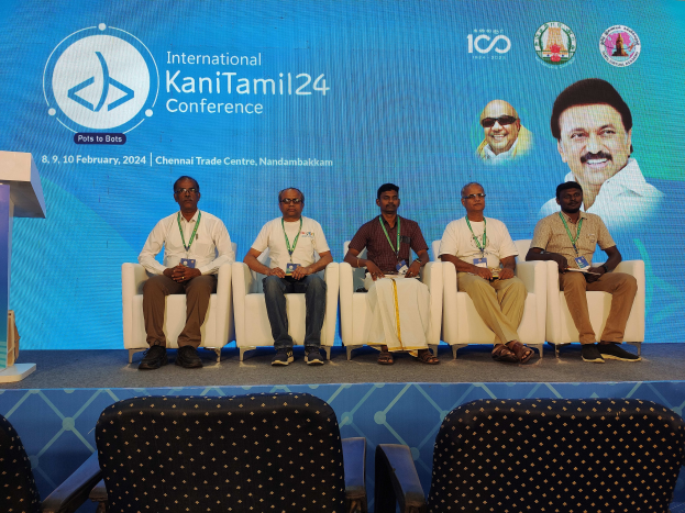 Eine Gruppe von Menschen, die vor einem Podium mit Mikrofon sitzen, mit einem Hintergrundbanner mit Text und Bildern von Menschen und Stühlen unten, das die Internationale Kanitamil24-Konferenz suggeriert.