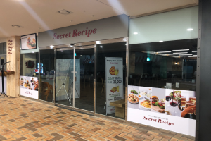 Der Eingang eines Restaurants namens "Geheimes Rezept" mit Glastüren, einem Stand mit verschiedenen Gegenständen und einer Speisekarte auf der linken Seite sowie Deckenlampen über den Türen.