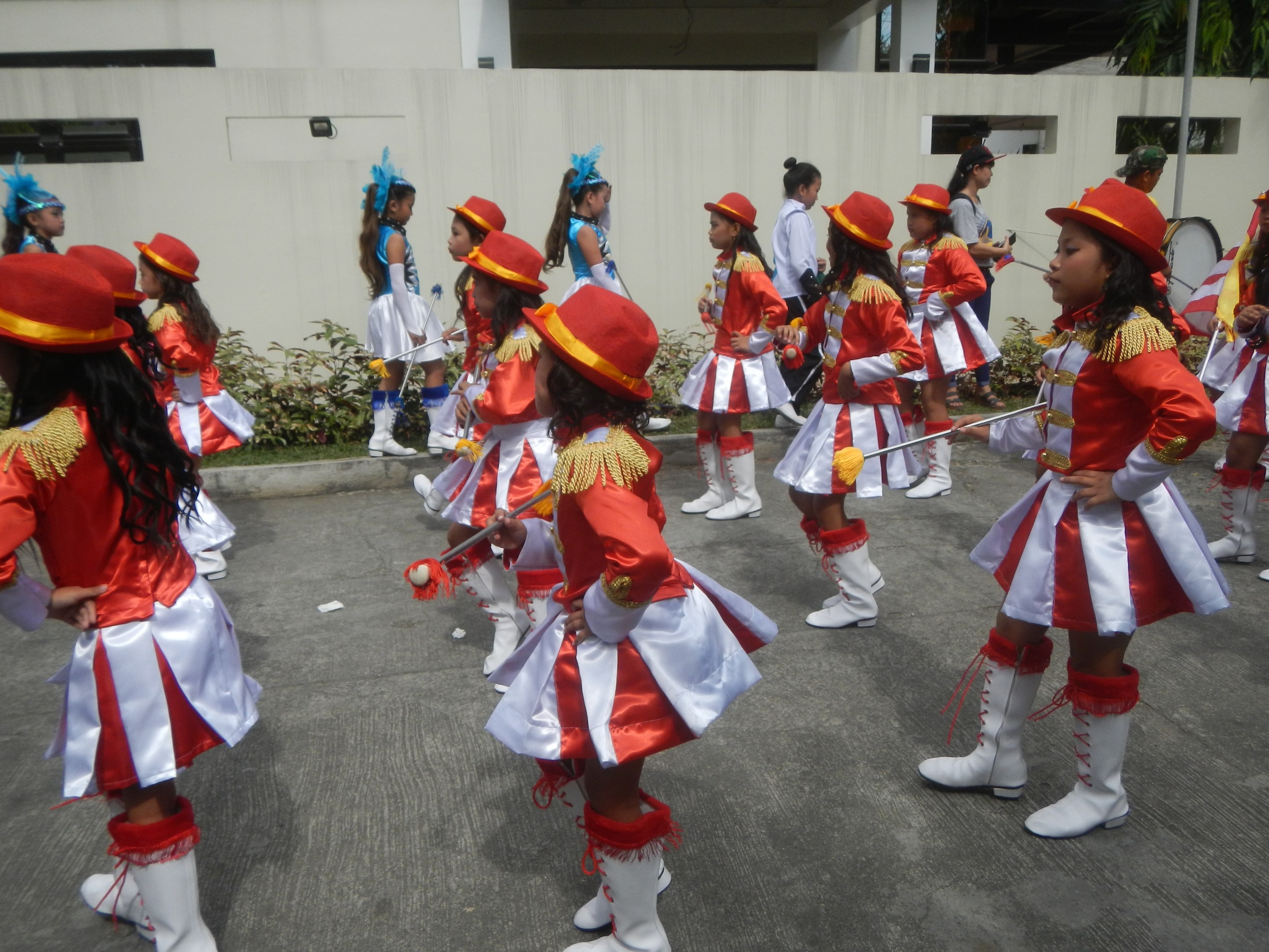 Gruppe junger Mädchen in roter und weißer Uniform, die marschieren und Musikinstrumente halten, mit einer Wand, Pflanzen, Bäumen und einem Gebäude im Hintergrund.