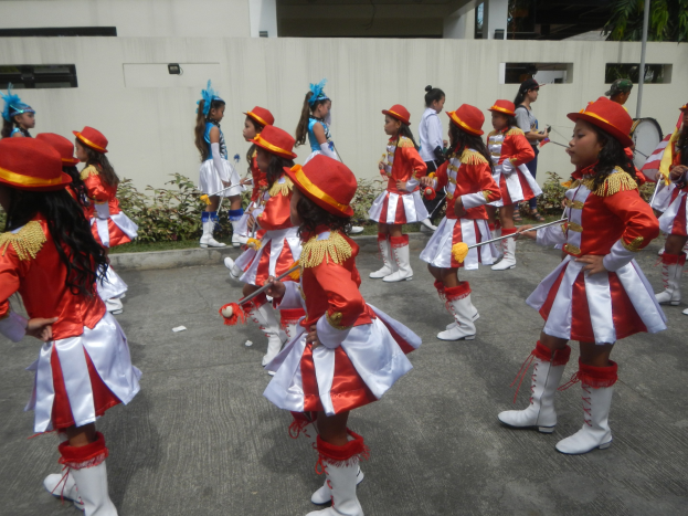 Gruppe junger Mädchen in roter und weißer Uniform, die marschieren und Musikinstrumente halten, mit einer Wand, Pflanzen, Bäumen und einem Gebäude im Hintergrund.