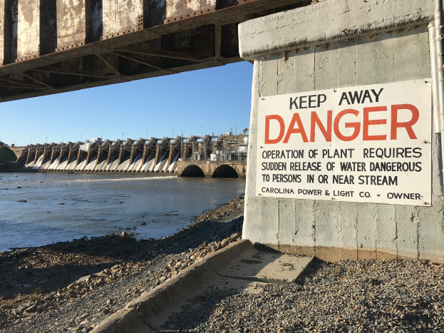 Eine Brücke mit einem "Achtung Gefahr" Schild, umgeben von Wasser, mit Gebäuden und einem klaren blauen Himmel im Hintergrund.