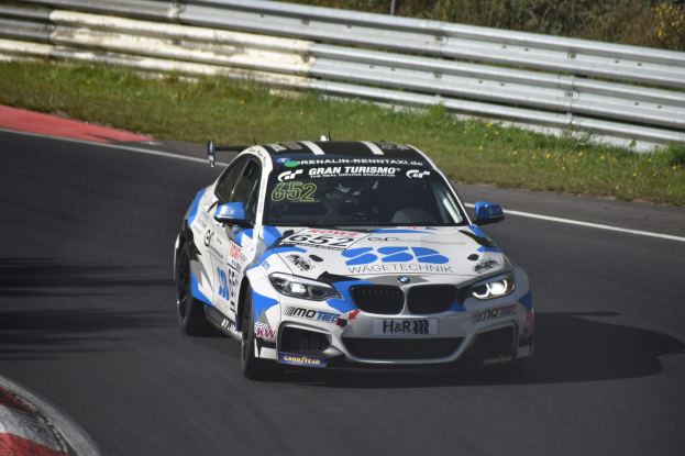Ein BMW M4 DTM-Rennwagen mit Text und Zahlen auf seiner Karosserie fährt auf einer Rennstrecke umgeben von Gras, einem Zaun und Pflanzen, wobei ein Rad sichtbar ist.
