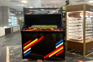 Ein Geschäft mit einem zentralen Retro-Arcade-Automaten, einem Gestell mit Gläsern auf der rechten Seite und Hintergrundelementen wie Schränken, einem Fernseher und anderen Gegenständen, mit Deckenleuchten, die eine Retro-Atmosphäre schaffen.