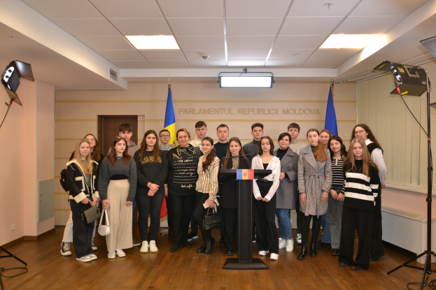 Eine Gruppe von Menschen vor einem Podium mit Mikrofon, umgeben von Fahnen, einer Wand mit dem Text "Moldova Youth Congress 2018", einer Tür, einem Dach mit Deckenleuchten und Ständern mit Leuchten.