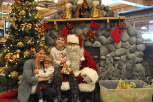 Eine Frau, Kinder und ein als Santa Claus verkleideter Mann mit Brille und Handschuhen stehen neben einem geschmückten Weihnachtsbaum, einer Wand mit Felsen, Spielzeugen, Dekorationen und einem Korb mit Gegenständen, mit Lichtern im Hintergrund.