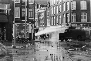 Schwarz-weiß-Foto einer Stadtstraße mit einem Feuerwehrauto, das Wasser versprüht, Gebäuden mit Fenstern im Hintergrund, einem Straßenschild, Menschen auf dem Gehweg und parkenden Fahrrädern.