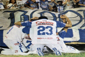 Eine Gruppe von Fussballspielern feiert ein Tor auf dem Boden vor einer jubelnden Menge, mit einem Banner mit Text im Hintergrund.