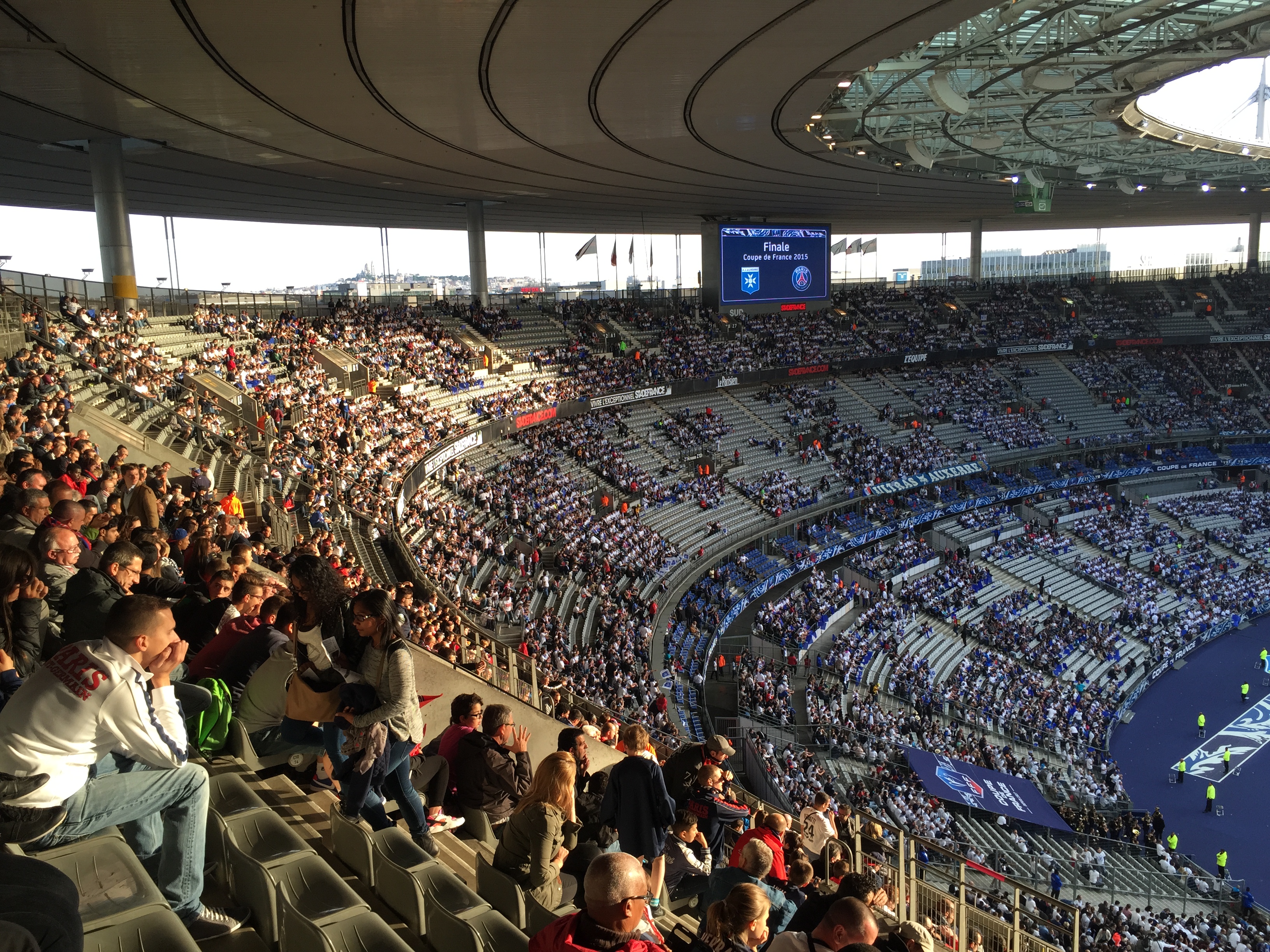 Eine große Menschenmenge sitzt im Allianz Stadion in München, Deutschland, und schaut ein Fußballspiel, mit einer Bühne auf der rechten Seite und Fahnen, Stangen und einem Bildschirm im Hintergrund.