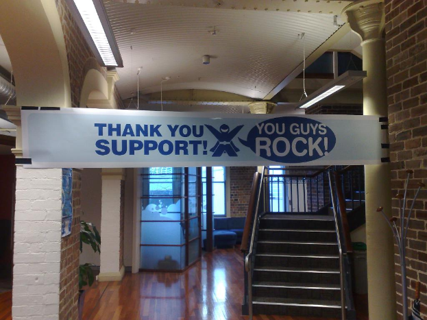 Dankeschön-Banner hängt an einer Wand und Säule vor einer Treppe, mit Fenstern im Hintergrund.