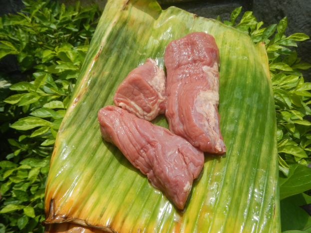 Drei Fleischstücke auf einem Bananenblatt, umgeben von grünen Blättern mit einer Wand im Hintergrund.