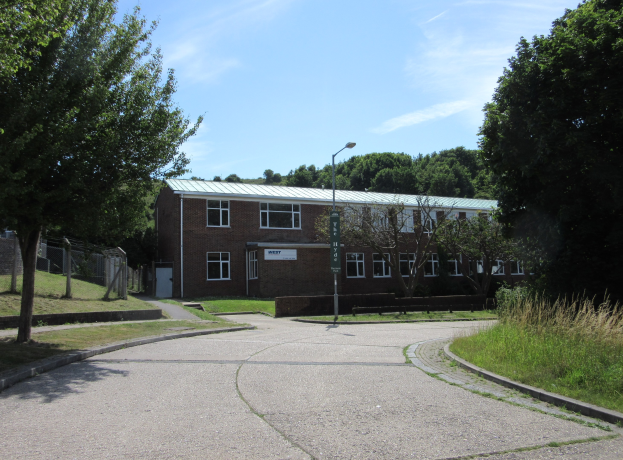 Ein Schulgebäude mit Fenstern, einem Straßenpfahl, einem Schild, einer Baumgruppe, Gras, einem Zaun und einem bewölkten Himmel, das in der Mitte eines Parks umgeben von saftigem Grün liegt.