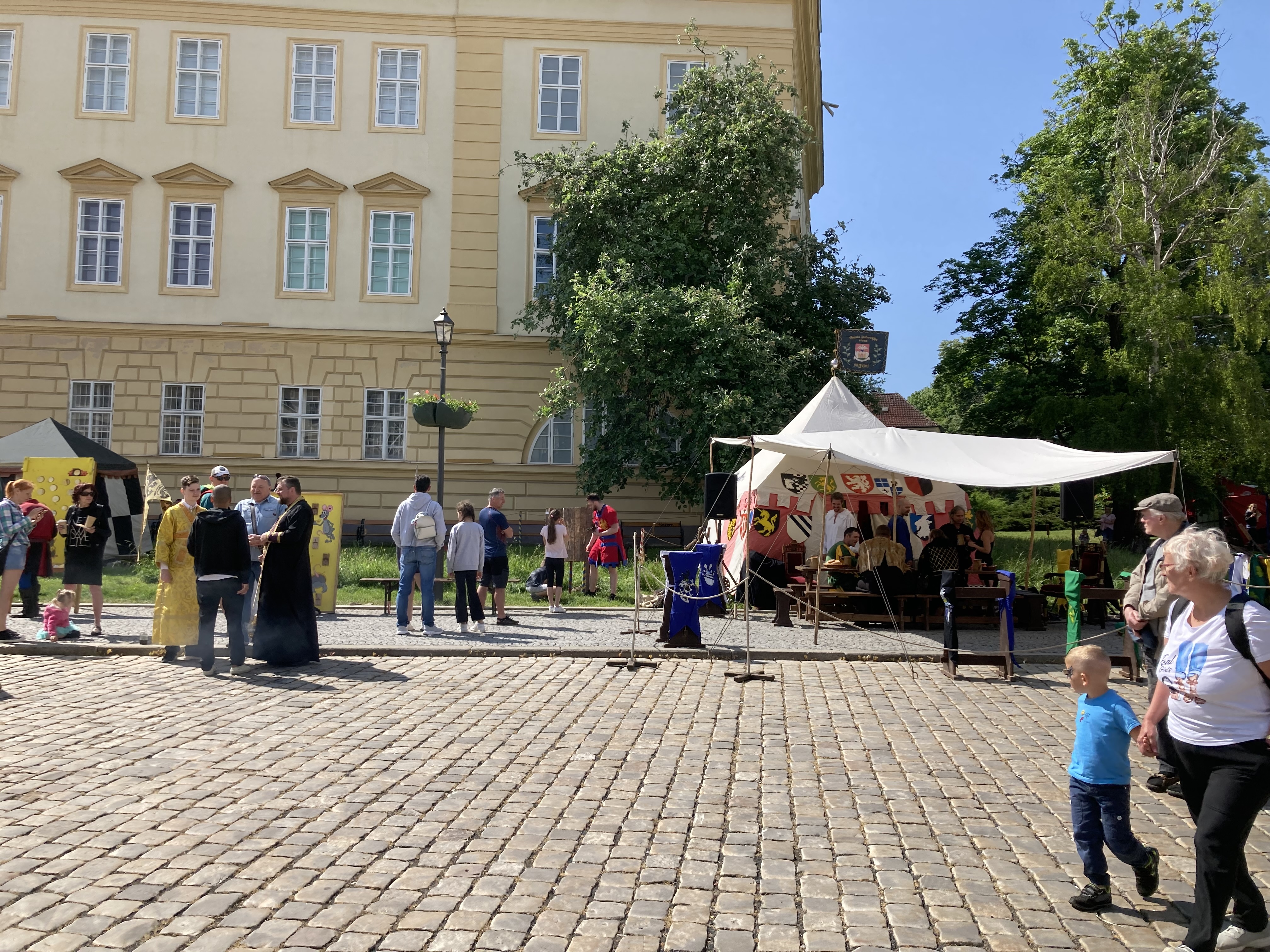 Eine Gruppe von Menschen geht eine Kopfsteinpflasterstraße in Krakau, Polen, entlang, mit Zelten, Tischen und Bäumen an den Seiten, vor einem Gebäude mit sichtbarem Text.
