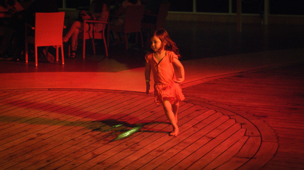 Ein Kind läuft auf einer hölzernen Plattform drinnen, mit ein paar Menschen im Hintergrund auf Stühlen sitzend.