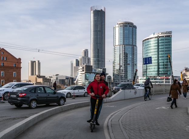 Ein Mann in einer roten Jacke und schwarzen Hosen fährt auf einem Elektroroller die Straße mit hohen Gebäuden hinunter, umgeben von Fußgängern und Radfahrern unter einem teilweise bewölkten Himmel.