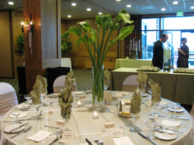 Ein Tisch mit einer weißen Decke, Tellern, Besteck, einem Glas und Zweigen, umgeben von Stühlen, mit einem grünen Tisch dahinter; ein Mann in einer schwarzen Jacke und eine Frau in der Nähe einer Tür mit zwei Männern dahinter.