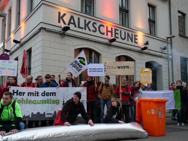 Eine Gruppe von Menschen mit Protestplakaten und Schildern vor einem Gebäude, mit zwei Personen im Vordergrund sitzend und einem Müllimer auf der rechten Seite; Gebäude mit Fenstern und Schildern im Hintergrund.